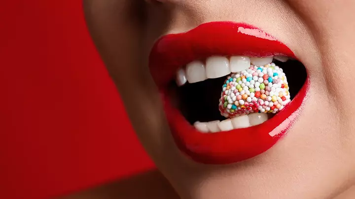 A closeup of a young woman's red lips and teeth biting into a colorful candy A closeup of a young woman's red lips and teeth biting into a colorful candy